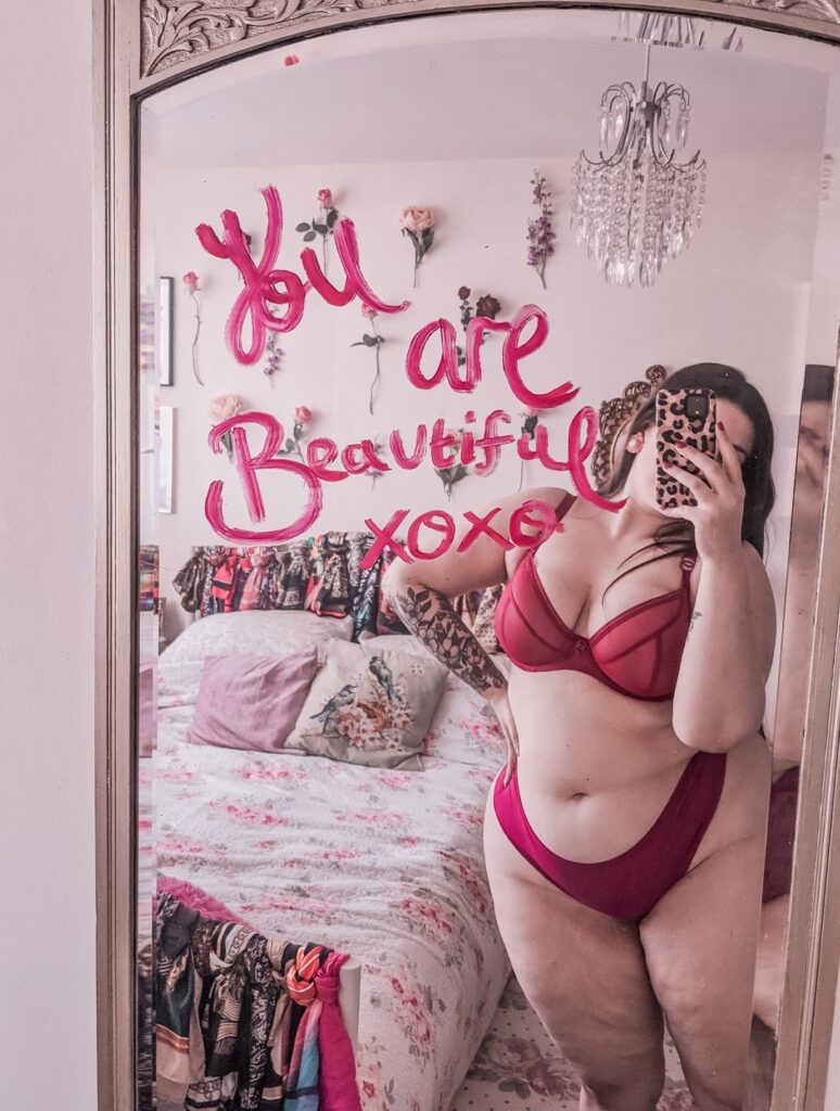 Mollie Quirk in pink matching underwear in front of a mirror that reads you are beautiful xoxo