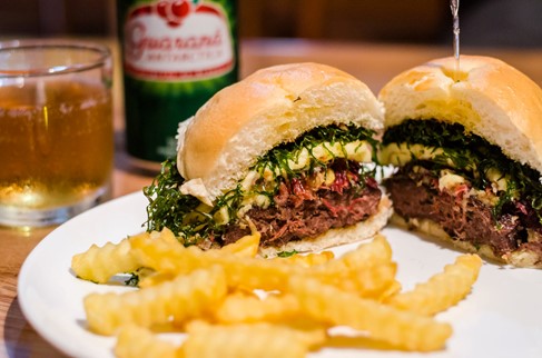 Photograph of a beef burger cut in half on a white plate with a side of chips and a blurred bottle of beer in the background