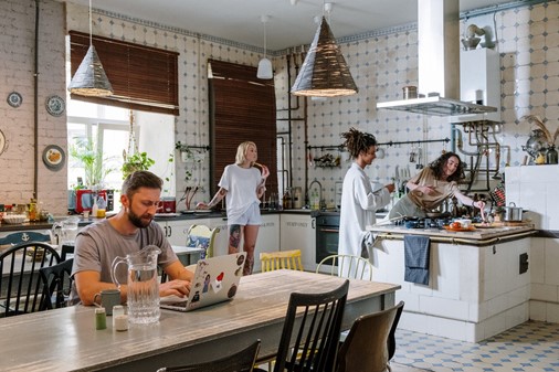 Photograph of four people in an open space kitchen one working on his laptop at a wooden table and the others eating food at the kitchen counter