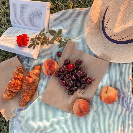 Summer fruits and croissants next to a half open book and sunhat on a soft white striped blanket