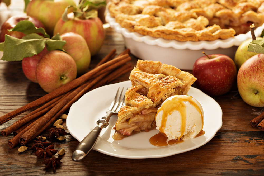 Photograph of a Caramel apple pie surrounded by fruit with the full pie in the background