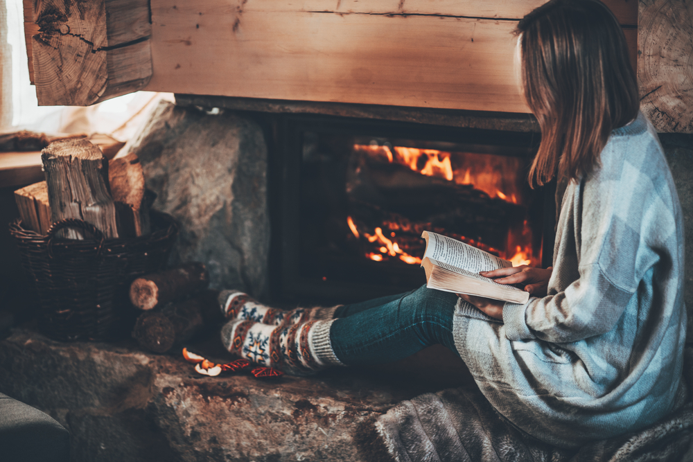 Photograph of a woman reading a book by a log burning fire
