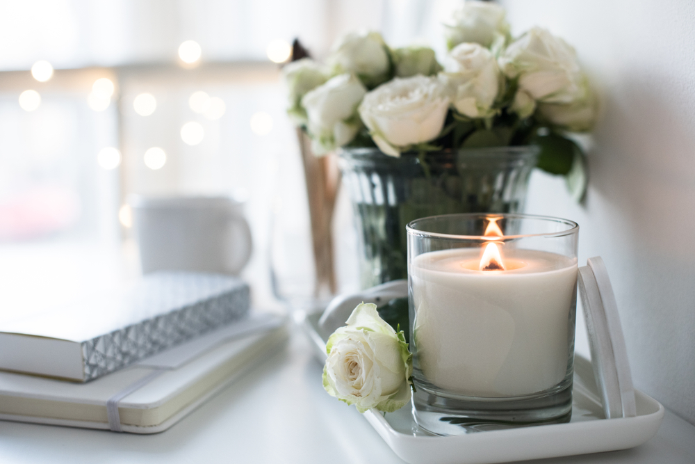 Image of a lit candle in a glass jar on a shelf next to some white roses and books