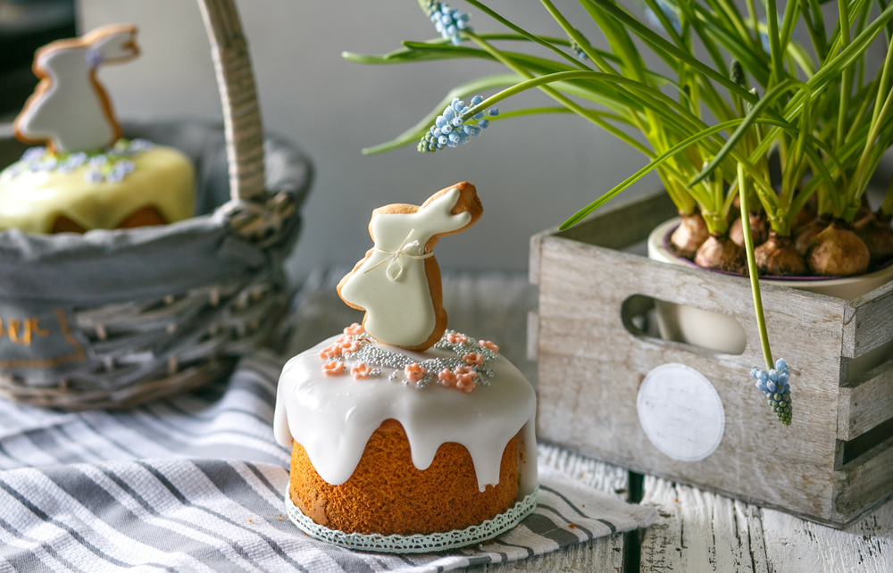 An easter cake with white icing  sprinkles and a rabbit topper on a table next to an easter basket and daffodils