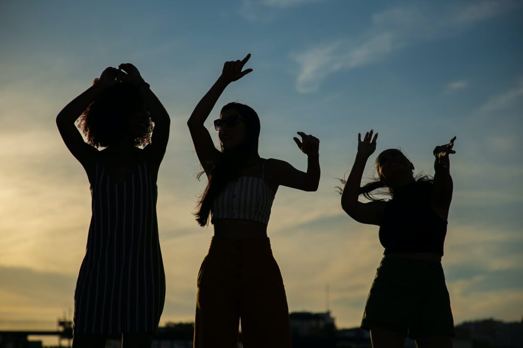 An image of three shadowed women dancing with their arms in the air against a clouded blue sky. 