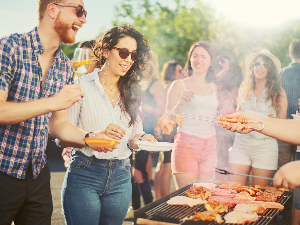 Group of family and friends at a barbeque being serviced bacon, sausages and beef patties