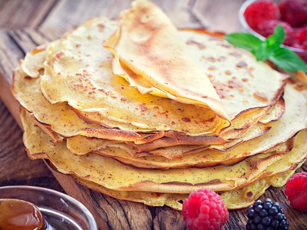 Homemade pancakes on a wooden board with a small pot of honey and summer fruits nearby