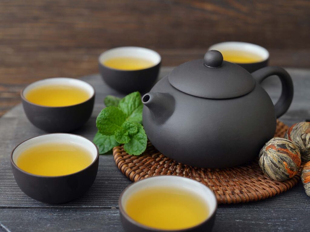 Matt black teapot on a wicker placemat with small matt black tea cups with healthy tea surrounding it