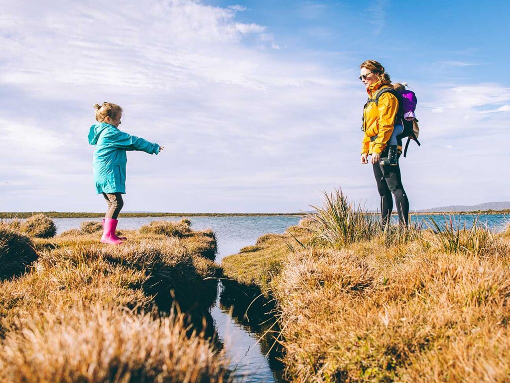 Mother and young daughter on a hiking journey next to a large lake standing on mounds of grass
