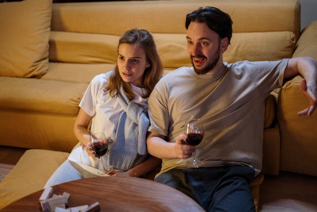 Photograph of a male and female couple sat leaning against a yellow couch holding glasses of red wine, shocked by something they see on TV. 