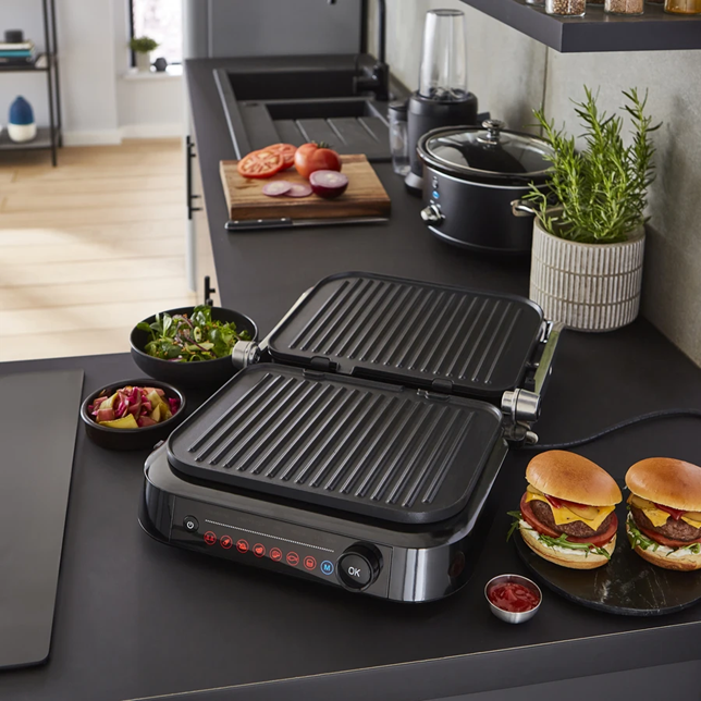 Photograph of the Swan Stealth Smart Grill open on a black countertop next to two burgers, side of coleslaw and salad