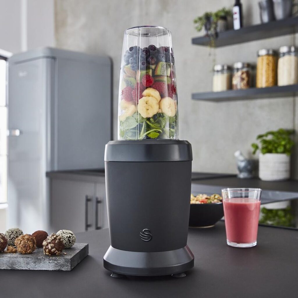 Photograph of the black Swan Stealth Personal Blender in a black fitted kitchen full with various fruits and spinach next to a plate of chocolate, a smoothie and a bowl of cereal.