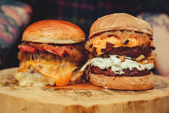 Photograph of two beef burgers with bacon and melted cheese resting on a wooden chopping board