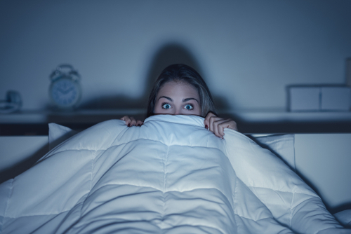 Photograph of woman in bed with her duvet covering her eyes watching a scary movie