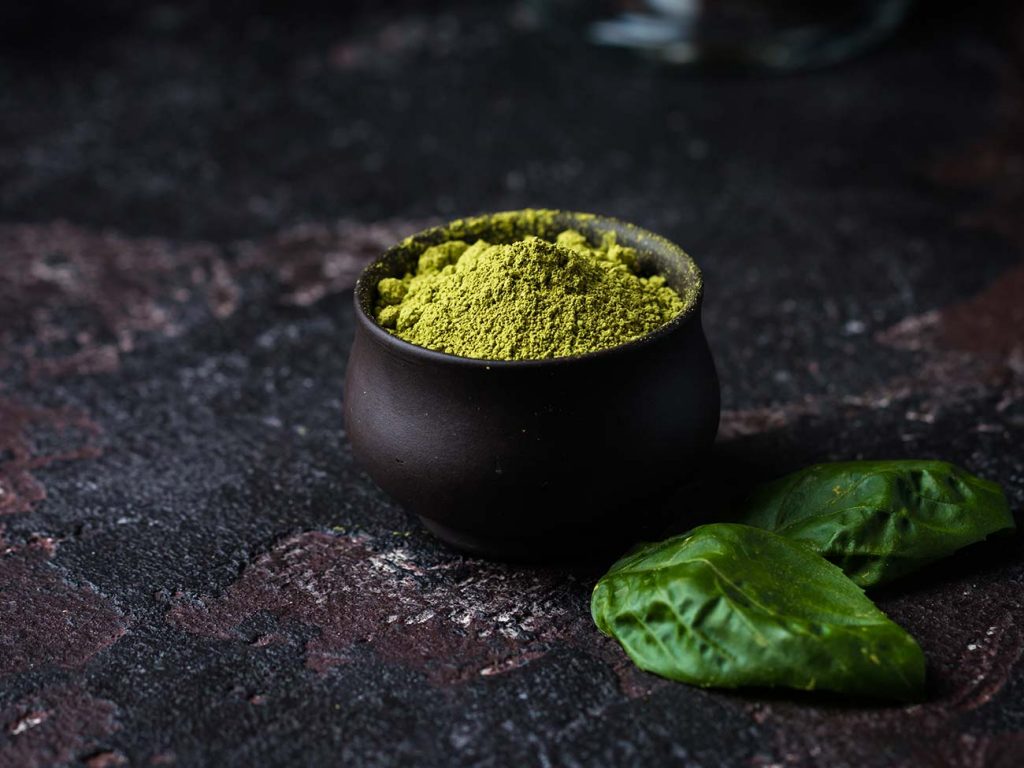 Small black pot of green matcha power next to matcha leaves against a dark stone background