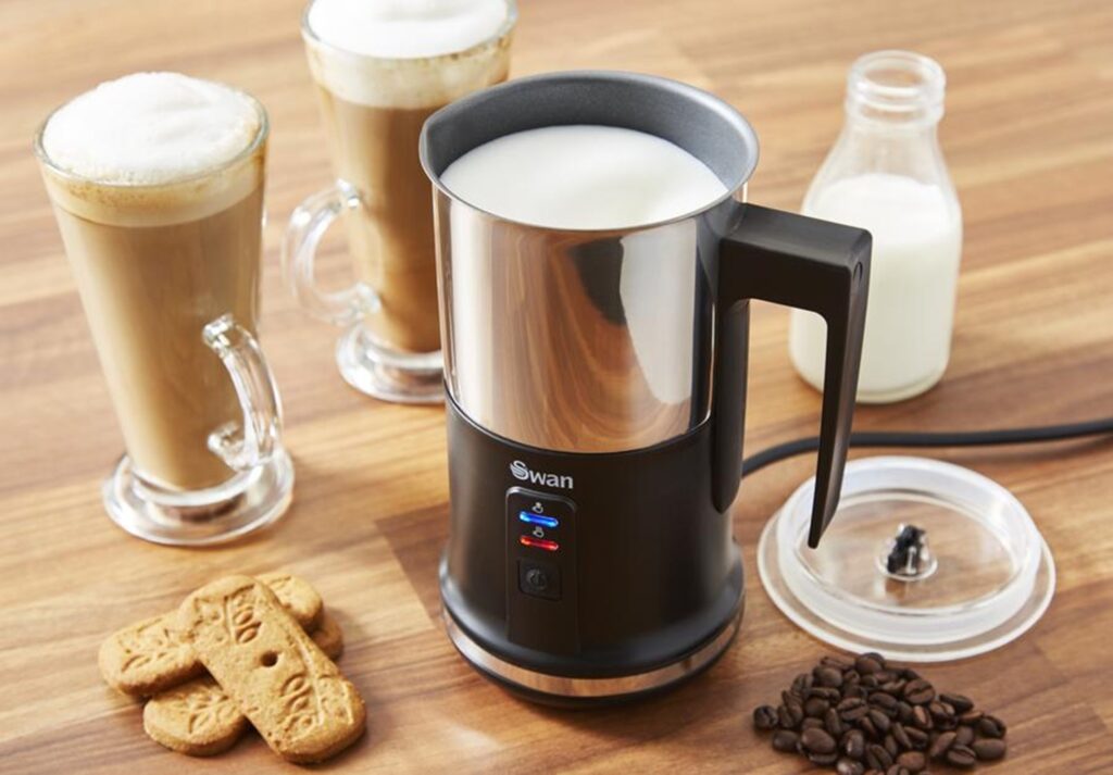 Swan Automatic Milk Frother next to two lattes, biscotti, a small jug of milk and a pile of espresso beans on a wooden countertop