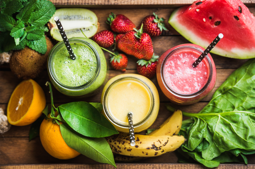 Three veganuary Fat-Burning Super Fruit Smoothie in colours green, yellow and red with fruits like bananas, strawberries and melon in the background