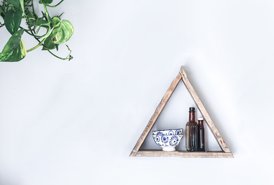 Triangle wooden shelf on a white wall holding two brown glass bottles and two blue and white patterned bowls