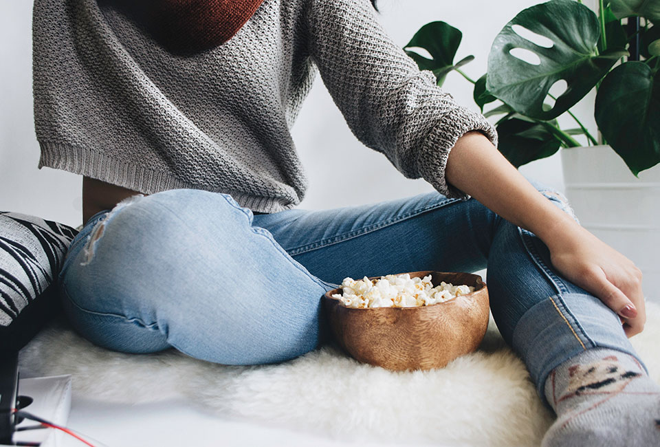 Woman in a grey crop sweatshirt and blue jeans eating popcorn from a small wooden bowl whilst sat on a white fur rug next to a potted green plant