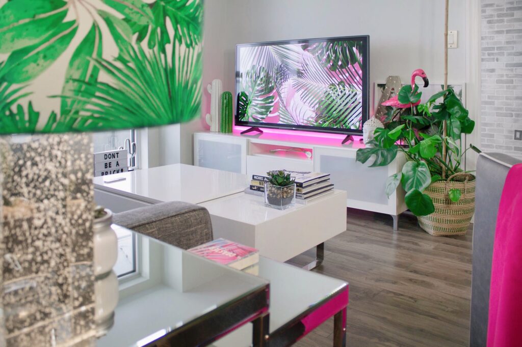 A picture of a living room decorated with tropical plants, prints and a flamingo. A TV screen with pink and green palm leaves on it is central to the image.