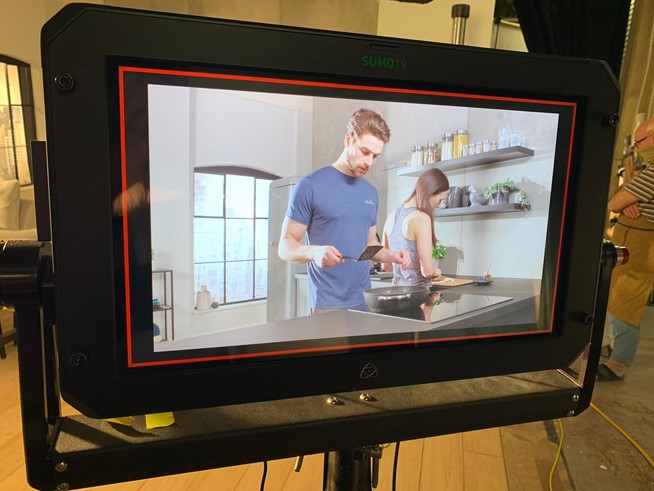 Close-up of a TV monitor with a man and female actor utilising the Swan Stealth Frying Pan on a fitted kitchen set