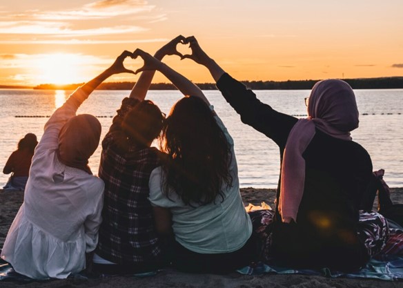 Group of women with their backs to the camera, sat on a beach watching the sunset over the ocean and using their hands to make heart shapes