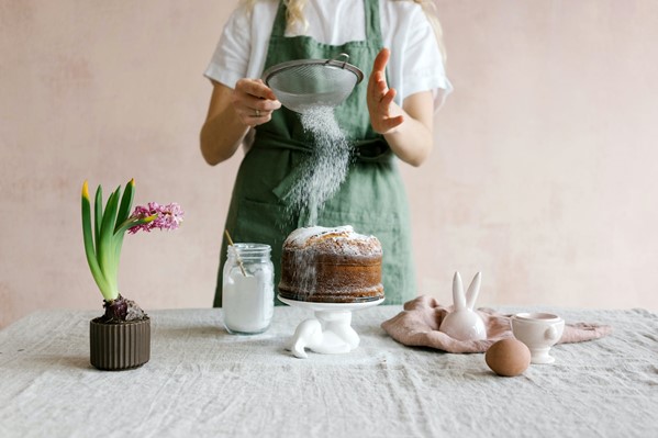 Shoulder down photograph of a woman dusting a sponge cake with icing sugar on a white table next to a potted blosso plan, an egg and a jar of flour