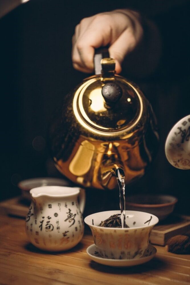 Image of a hand pouring a cup of tea out of a golden tea pot into a mug full of tea leaves with gold chinese writing around the outside, in the background is a matching milk jug