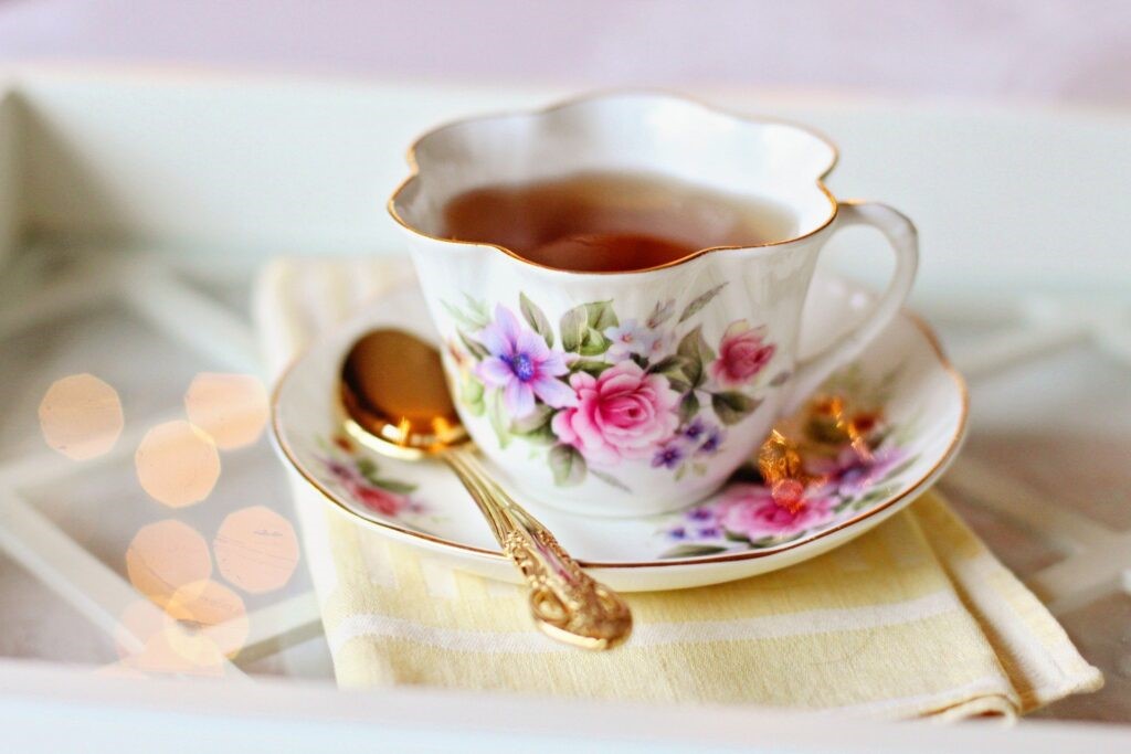 Picture of a white teacup and saucer with a pink and purple floral pattern, filled with tea. On the saucer is a gold teaspoon, matching the gold rim of the tea cup
