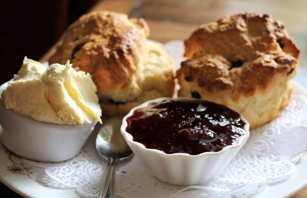 Image of two scones on a plate with a pot of clotted cream and a pot of jam next to a silver teaspoon