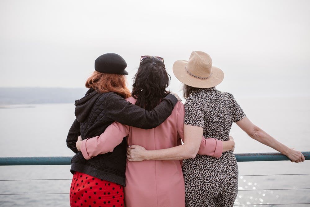 Three women with their backs turned to us with their arms around each other looking out towards the sea and sky.