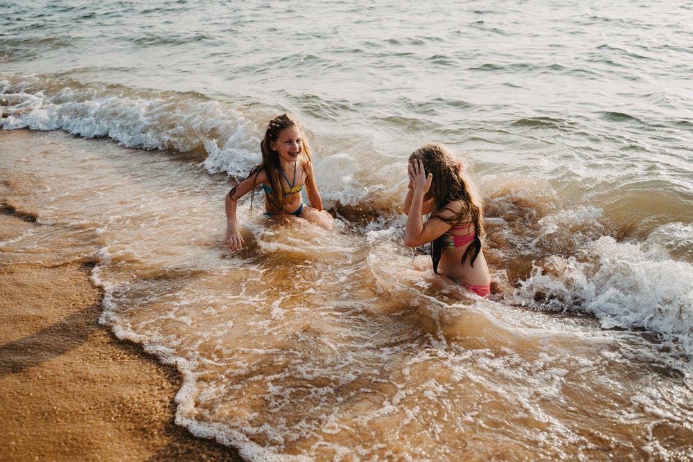Two small female children with long hair, sitting down in the shallow waves of the beach. Laughing and playing.