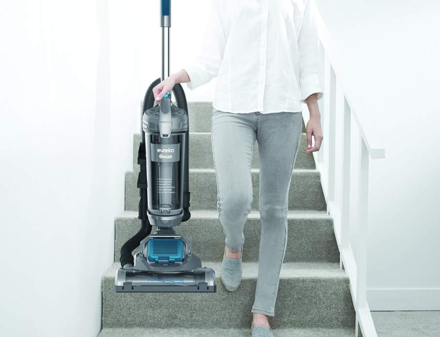 Woman walking down some grey carpeted stairs in a white shirt and light grey jeans with Vacuum in one hand.