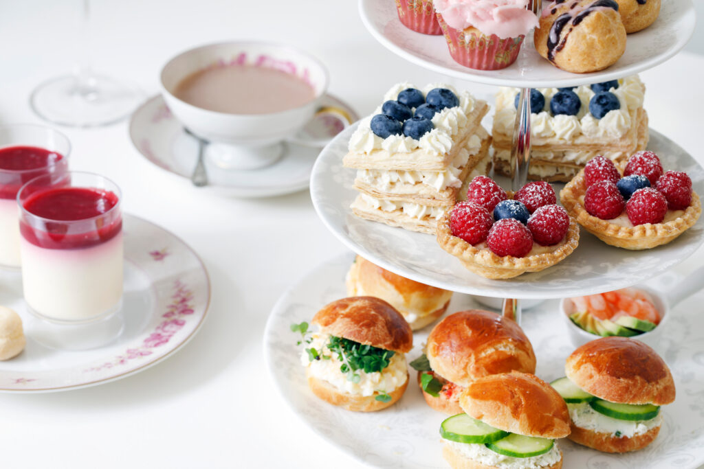 Image of an afternoon tea with mini brioche canapes and a selection of sweets and cakes on a tiered stand. In the background are a cup of tea in a cup and saucer and a plate with more desserts.