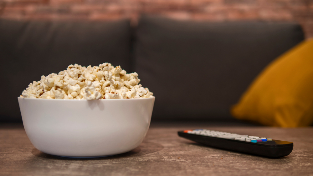 A white bowl filled with popcorn on a wooden table, next to a remote. 