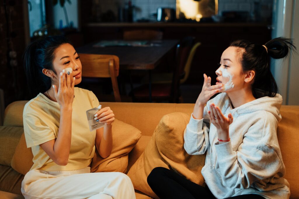 Two women sat on an orange sofa, applying a white face mask, one wearing a t-shirt and one wearing a hoodie.