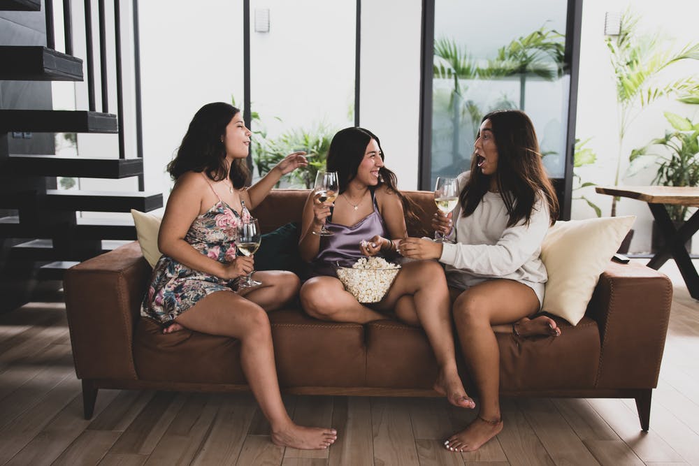 Three women sat on a brown sofa in a brightly lit living room, sharing popcorn and drinking white wine.