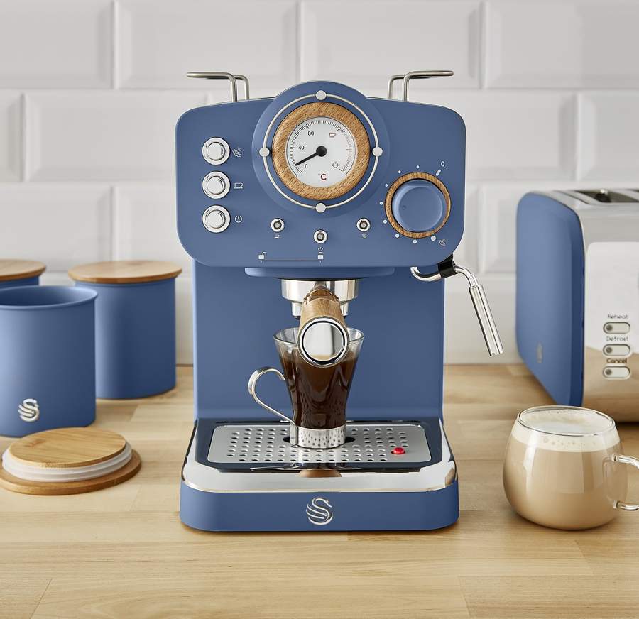 A Swan branded Espresso Machine in dark navy blue pouring a black coffee with a latte coffee next to it. The coffee machine sits between a nordic style toaster and canisters to match.