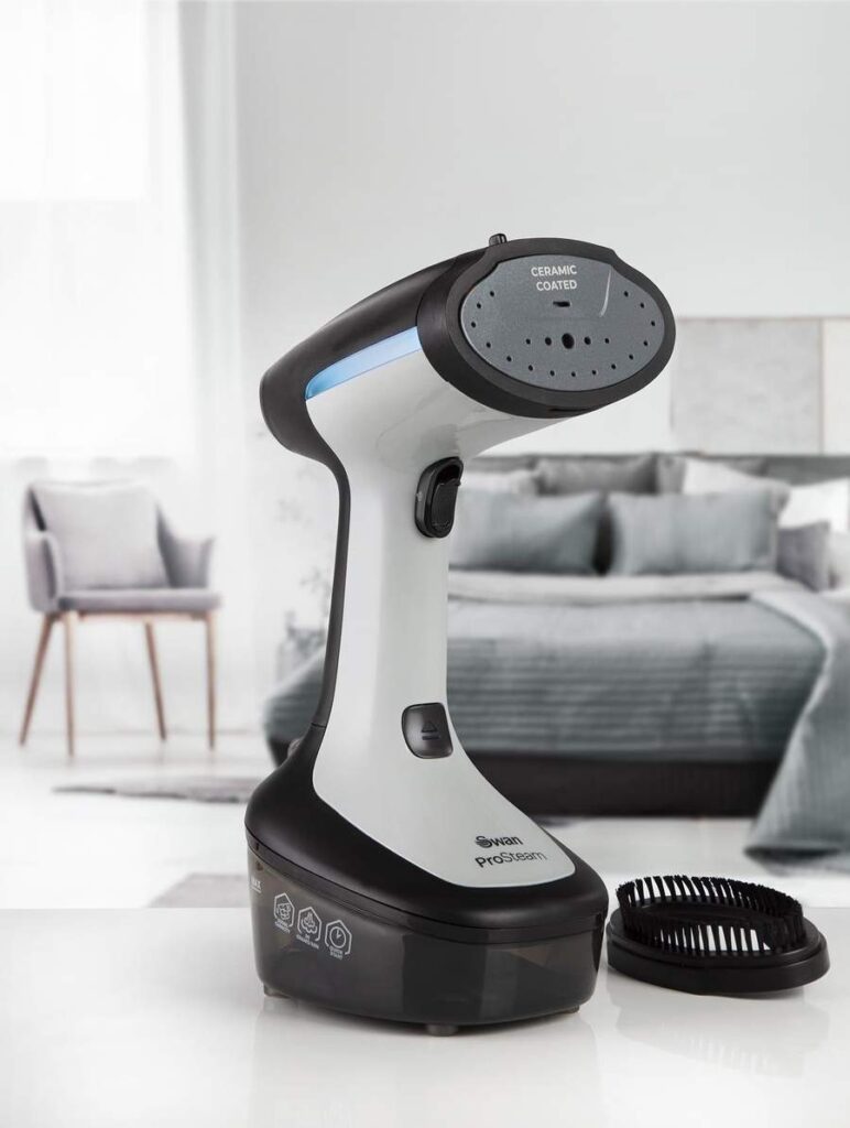 Black and white garment steamer on a desk with grey curtains, bedding and cushions in the background.