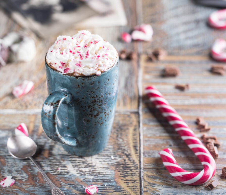 A wooden table with a candy cane resting on the table with a cup of peppermint white hot chocolate resting next to the cane on a christmas dinner recipes blog.