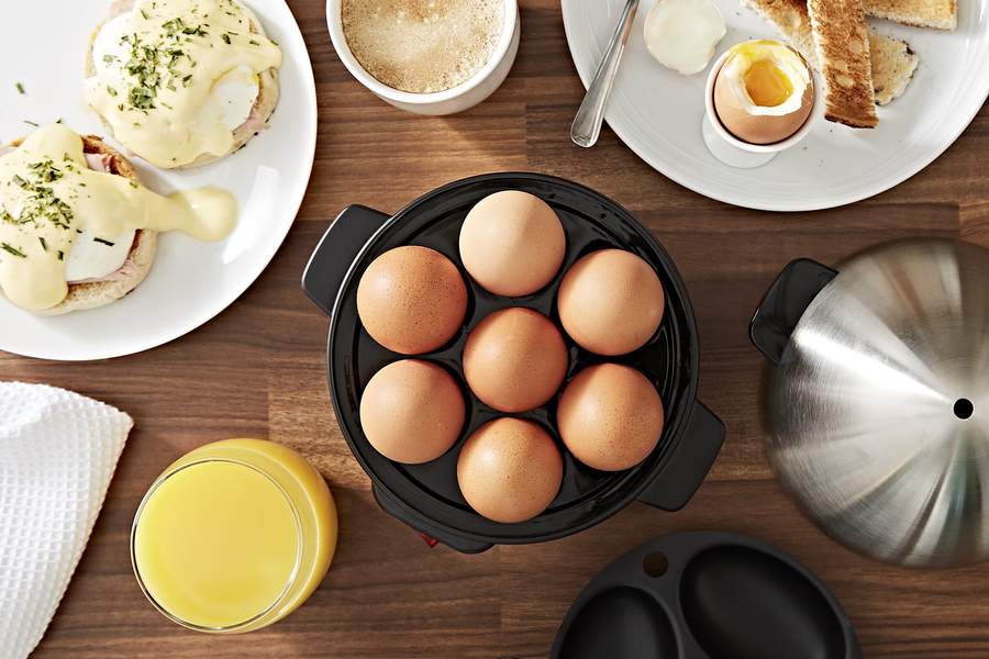 An overhead view of an egg boiler filled with boiled eggs, surrounded by plates of egg dishes and cups of coffee/orange juice.
