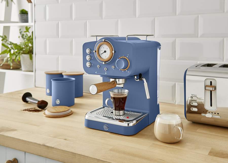 A dark blue Nordic Espresso Coffee Machine on a light wood countertop next to glasses of coffee, a dark blue nordic toaster, and canister set.