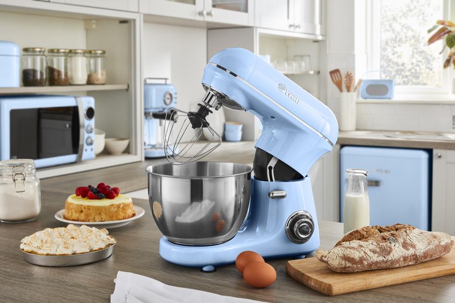 A Blue Retro Stand Mixer on a kitchen counter top surrounded by baked cakes and bread, More Blue Retro kitchen items in the background to match.