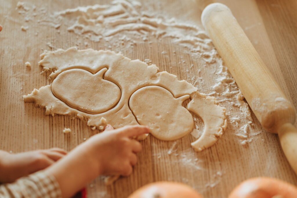 A chopping board with dough and a rolling pin displayed to show two pumpkin cut-outs in the dough. A child's hands are reaching for the excess dough.