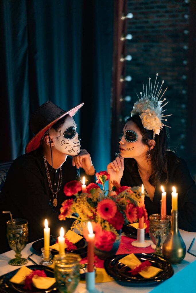 A couple sat at a dark dinner party looking at each other. Both of their faces are decorated in Halloween face paints. The table is filled with flowers and multicoloured (lit) candles.
