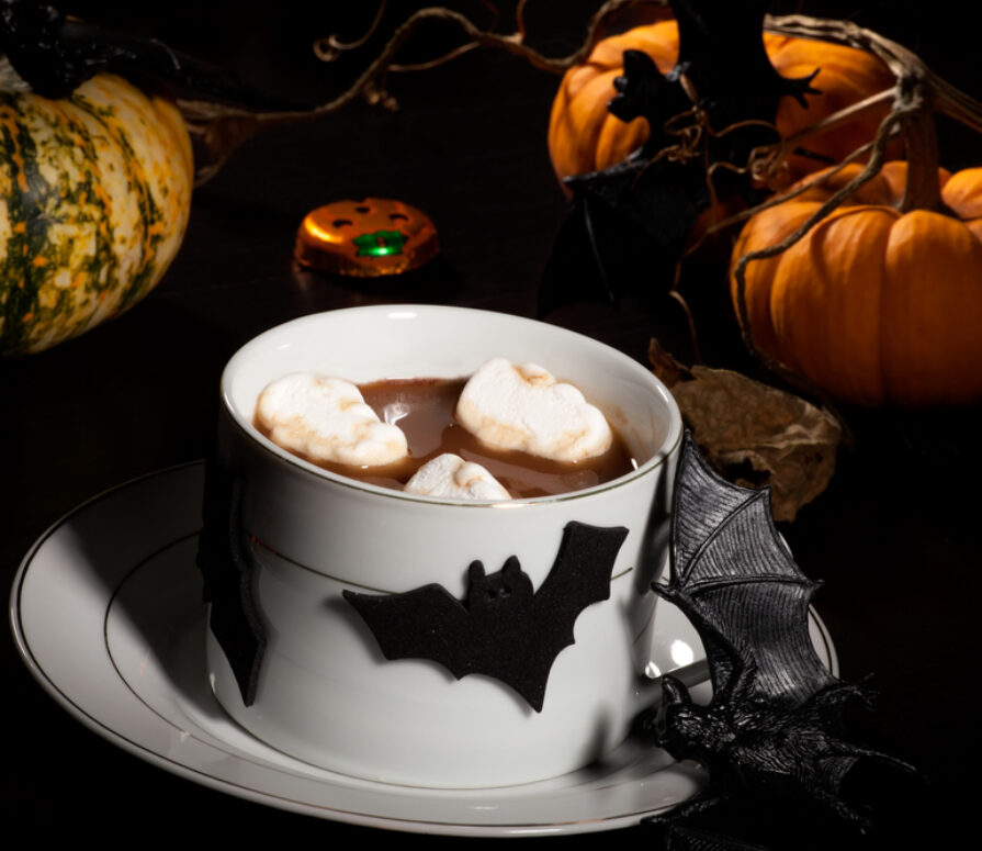 A white tea cup and saucer full of hot chocolate and marshmallows, in front of some pumpkins and decorated with Halloween decor.