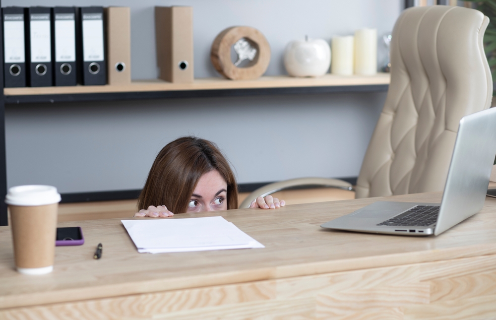 Young female office worker hiding behind desk and looking at laptop