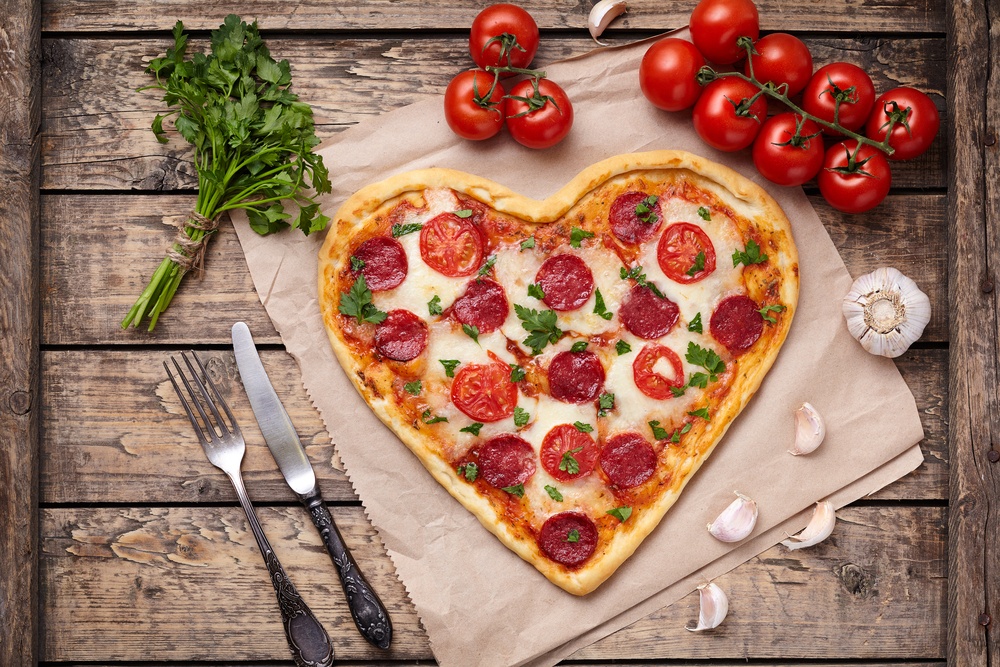Heart shaped pepperoni pizza next to fresh tomatoes and basil