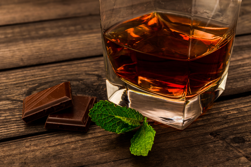 Short glass of whisky next to two pieces of dark chocolate and some mint
