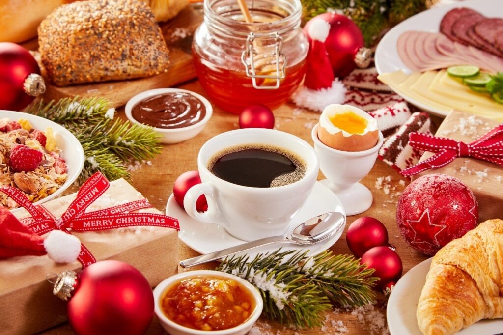 Image of a boxing day/Christmas day breakfast with a cup of coffee, a boiled egg, croissants, bread, granola, meats and cheeses. Scattered in between the food are presents, baubles, pine tree sprigs and snow. 